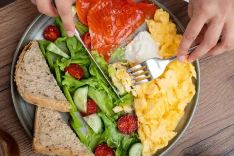 Assiette colorée avec saumon fumé, œufs brouillés, salade verte, tomates séchées, concombre et pain complet. Un repas protéiné parfait pour bien commencer la journée.