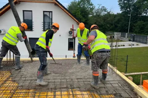 Budowlańcy w kamizelkach i kaskach, przestrzegając zasady bhp, wylewają beton. Jeden kieruje rurę, drugi grabi, trzeci wyrównuje.