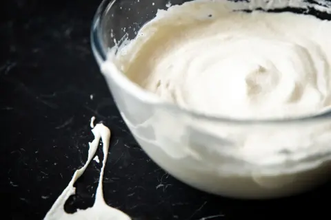 Schlagrahm in einer Glasschüssel, bereit für ein Dessert. Ein Klecks Sahne ist daneben auf der dunklen Arbeitsfläche zu sehen.