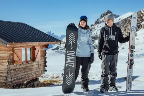 Para trzymająca deski snowboardowe przed drewnianą chatką w górach.