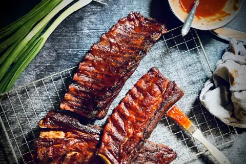 Saftige spare ribs glänzen in BBQ-Sauce auf einem Grillrost. Frühlingszwiebeln und eine Sauce-Schale deuten auf ein köstliches Mahl hin.