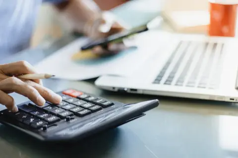 Dłoń z ołówkiem liczy na kalkulatorze, obok laptop i kubek. Analiza rachunków, może zużycie kwh.