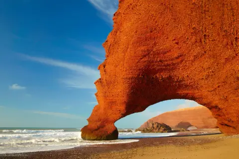 Pomarańczowe skały na plaży w Maroku, fale oceanu i błękitne niebo. Niesamowite widoki, a ceny w Maroku zachęcają do podróży.