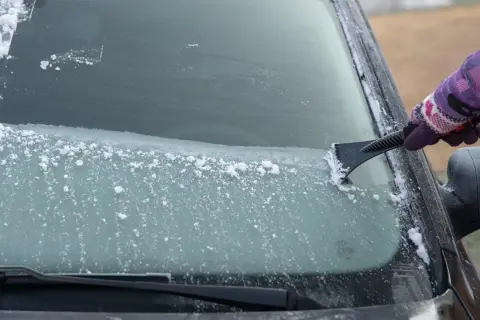 Ręka w rękawiczce zeskrobuje szron na szybie samochodu. Zima daje się we znaki, ale widok czystej szyby jest nagrodą.