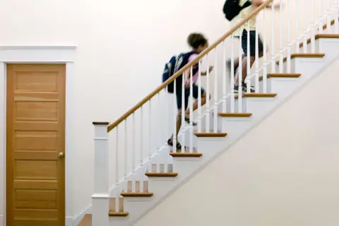 Two people with backpacks ascend a staircase with a new white stair railing and wooden handrail.