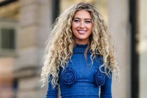 Mujer sonriente con un espectacular pelo chino rubio, luciendo un vestido vaquero azul con detalles de rosas.