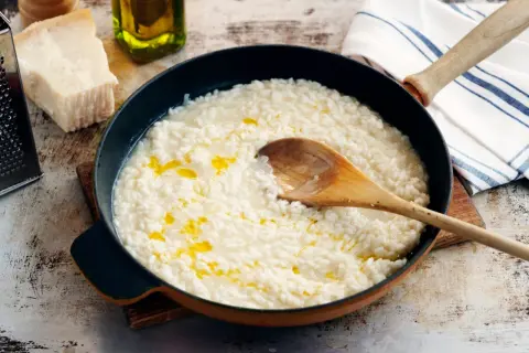 Krämig parmesanrisotto i en panna, med en träsked som rör om. Ingredienser som parmesanost och olivolja syns i bakgrunden.