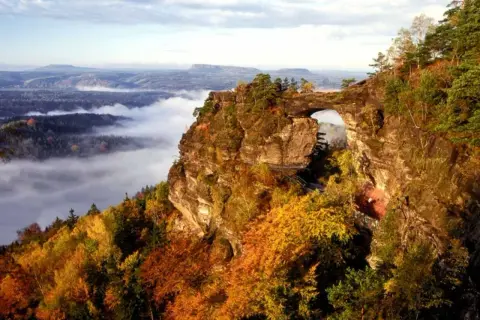Majestätische Felsformation Pravcicka brana mit einem natürlichen Torbogen, umgeben von herbstlichem Laub und Nebel im Tal.