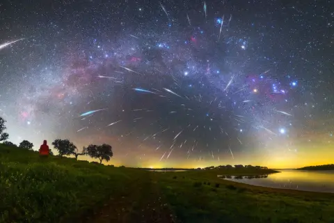 A lone figure watches a spectacular display of shoot stars streaking across the Milky Way above a tranquil lake and grassy hills.