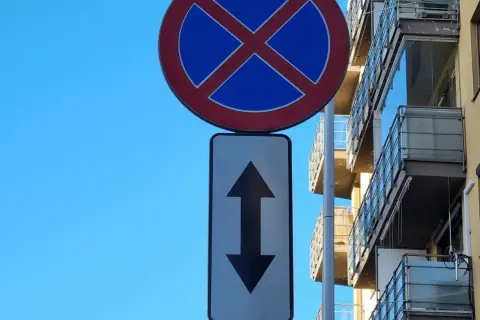 Znak zakaz postoju z tabliczką wskazującą, że zakaz obowiązuje na odcinku w obu kierunkach. Budynek z balkonami w tle.