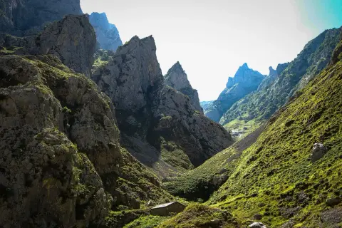 Kamienna chata w dolinie u stóp skalistych szczytów, przypominających kształtem **Naranjo de Bulnes**.
