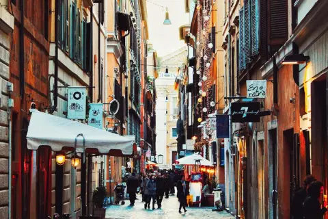 En livlig gränd i ett kvarter i Rom, med butiker och restauranger längs sidorna. Folk promenerar och njuter av atmosfären.