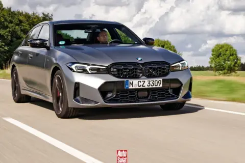 Szare BMW 320d jedzie po drodze, za kierownicą siedzi mężczyzna. W tle drzewa i chmury.