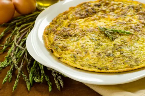 Frittata di asparagi selvatici dorata su piatto bianco, con asparagi freschi e uova sullo sfondo.