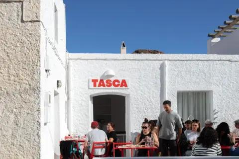 Gente disfrutando en el bar Tasca, con sus altas hortichuelas, bajo un cielo azul.