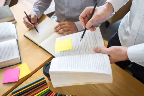 Dwie osoby uczące się przy biurku, otwarte książki, notatki i kredki.