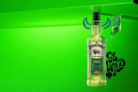 Butelka Żubrówki z trawą żubrową, 37,5% alkoholu. Obok kieliszek z zielonym płynem i napis "Go Wild".