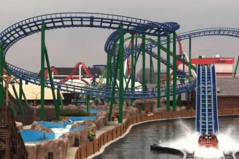 Water coaster zjeżdża z dużej wysokości, tworząc fontannę wody. W tle widać inne atrakcje parku rozrywki.