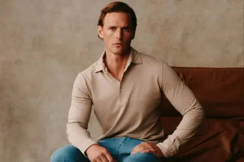 Ein Mann sitzt lässig in einem beigen Langarm-Polohemd, das an italienische Poloshirts für Herren erinnert.