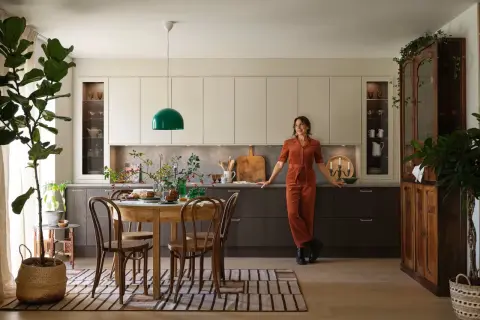 En kvinna i orange jumpsuit lutar sig mot köksbänken i ett kök med fransk inredningsstil. Ett runt träbord med stolar står framför köksöarna.