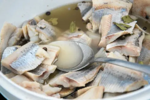 Kawałki śledzi w zalewie z przyprawami, gotowe do podania. Widać łyżkę zanurzoną w marynacie.