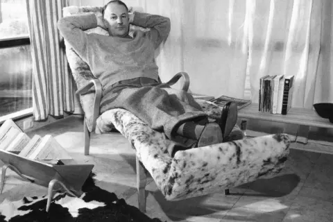 Man vilar i en Bruno Mathsson liggfåtölj, klädd i fårskinn. Böcker och tidningar ligger utspridda.