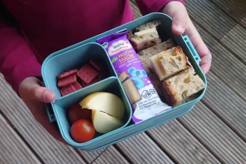 Proviant für die Wanderung: Brotscheiben mit Käse, rote Kekse, Apfelspalten und Tomaten in einer Brotdose. Ein Fruchtriegel liegt daneben.