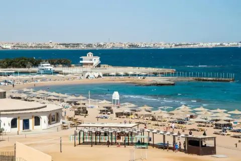 Solig strand i Hurghada, Egypten, med parasoller, solstolar och turkost vatten. En båt ligger vid en pir.