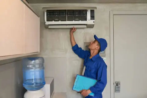Un technicien en uniforme bleu vérifie une installation de climatisation murale, tenant un dossier de service.