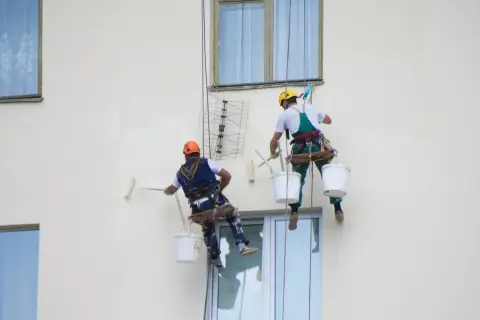 Deux peintres suspendus par des cordes peignent un mur extérieur. L'un utilise un rouleau, l'autre est prêt à commencer.