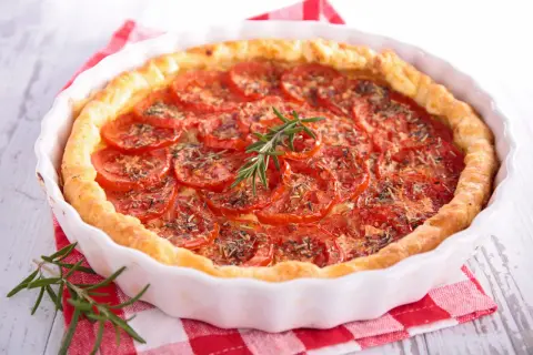 Une délicieuse tarte à la tomate, garnie de fines herbes et d'une branche de romarin frais, repose dans un plat blanc sur un torchon à carreaux rouges.