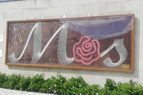 Letras de encaje blanco y rosa forman "Mos" en un letrero en el Pazo de Mos, con herramientas de encaje a la izquierda.