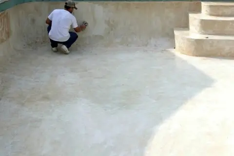 Hombre lijando la pared de una piscina vacía, preparándola para pintar cemento exterior.