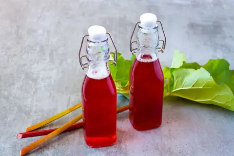 Dwie butelki z rubinowym sokiem z rabarbaru, obok łodygi i liście rabarbaru.