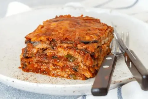 Porción de pastel de berenjenas, con capas de berenjena y salsa de tomate, servido en un plato blanco con cubiertos. ¡Un pastel de berenjenas facilisimo!
