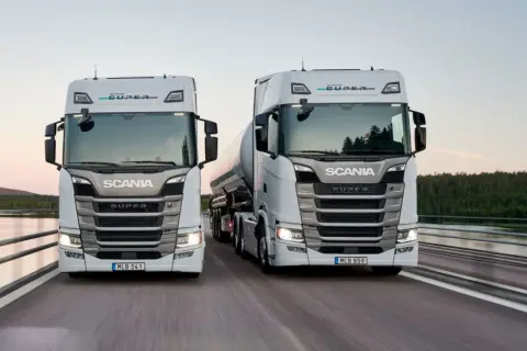 Två nya Scania lastbilar, en med tank, kör på en bro. Kolla in ny Scania lastbil pris för din nästa transport.