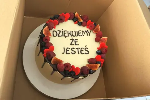 Okrągły tort z białym kremem, ozdobiony owocami i serduszkami. Krótki napis na tort urodzinowy: "DZIĘKUJEMY ŻE JESTEŚ".