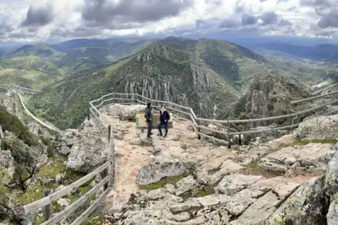 Dos personas admiran el paisaje montañoso desde el mirador del Cerro del Viso. El cielo está nublado.