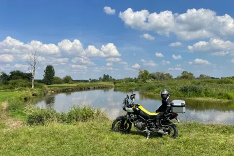 Motocyklista w kasku siedzi na motocyklu obok rzeki, na tle zielonej łąki i błękitnego nieba z chmurami.