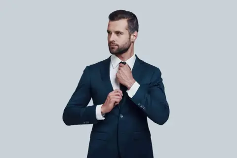 Elegancki mężczyzna w granatowym garniturze i białej koszuli poprawia krawat. Idealna stylizacja, gdy zastanawiasz się, jaka koszula do granatowego garnituru.