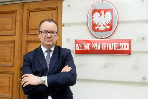 Mężczyzna w garniturze stoi przed budynkiem z godłem Polski i tabliczką "Rzecznik Praw Obywatelskich".