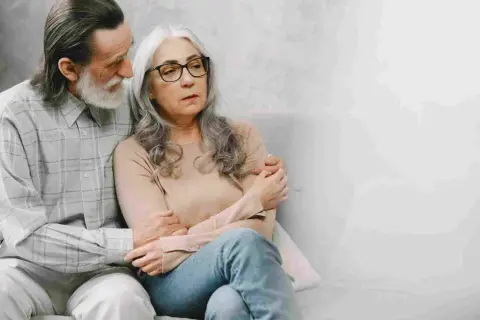 Coppia anziana seduta sul divano. Lui la conforta, forse parlando di come la pensione di invalidità è reversibile.