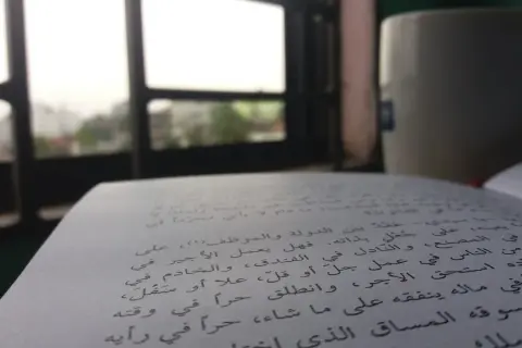 Otwarta księga z tekstem w języku arabskim, obok kubek i widok przez okno.