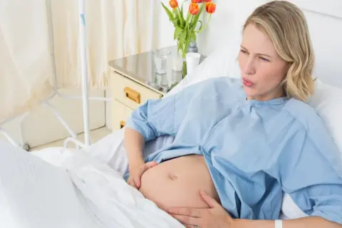 Kobieta w szpitalnym łóżku, z bólem na twarzy, trzymająca się za brzuch. Zastanawia się, jak wywołać poród.