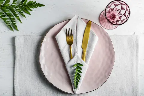 Service de table élégant avec pliage serviette communion, assiette rose, couverts dorés et verre à facettes.