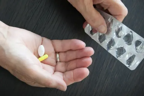 Dłoń trzyma białą i żółtą tabletkę. Obok puste opakowanie po lekach. Glicynian magnezu na co? Na stres i zmęczenie.