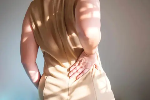 Une femme se tient le bas du dos, ressentant une lourdeur. La lumière du soleil projette des ombres sur sa peau.