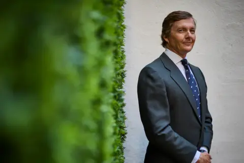 Cayetano de Borbón-Dos Sicilias, con traje gris y corbata azul, posa con una sonrisa ante una pared blanca y vegetación verde.
