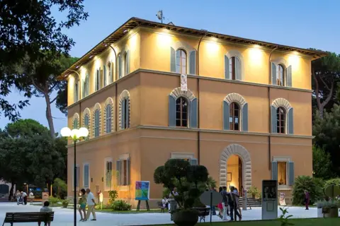 Edificio giallo illuminato nel parco della Versiliana, con persone che passeggiano e un'opera d'arte esposta.