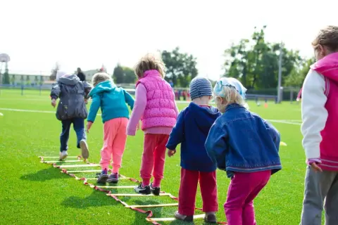Niños corriendo en una escalera de agilidad en un campo de césped artificial, ideal para aprender inglés en lugares divertidos.